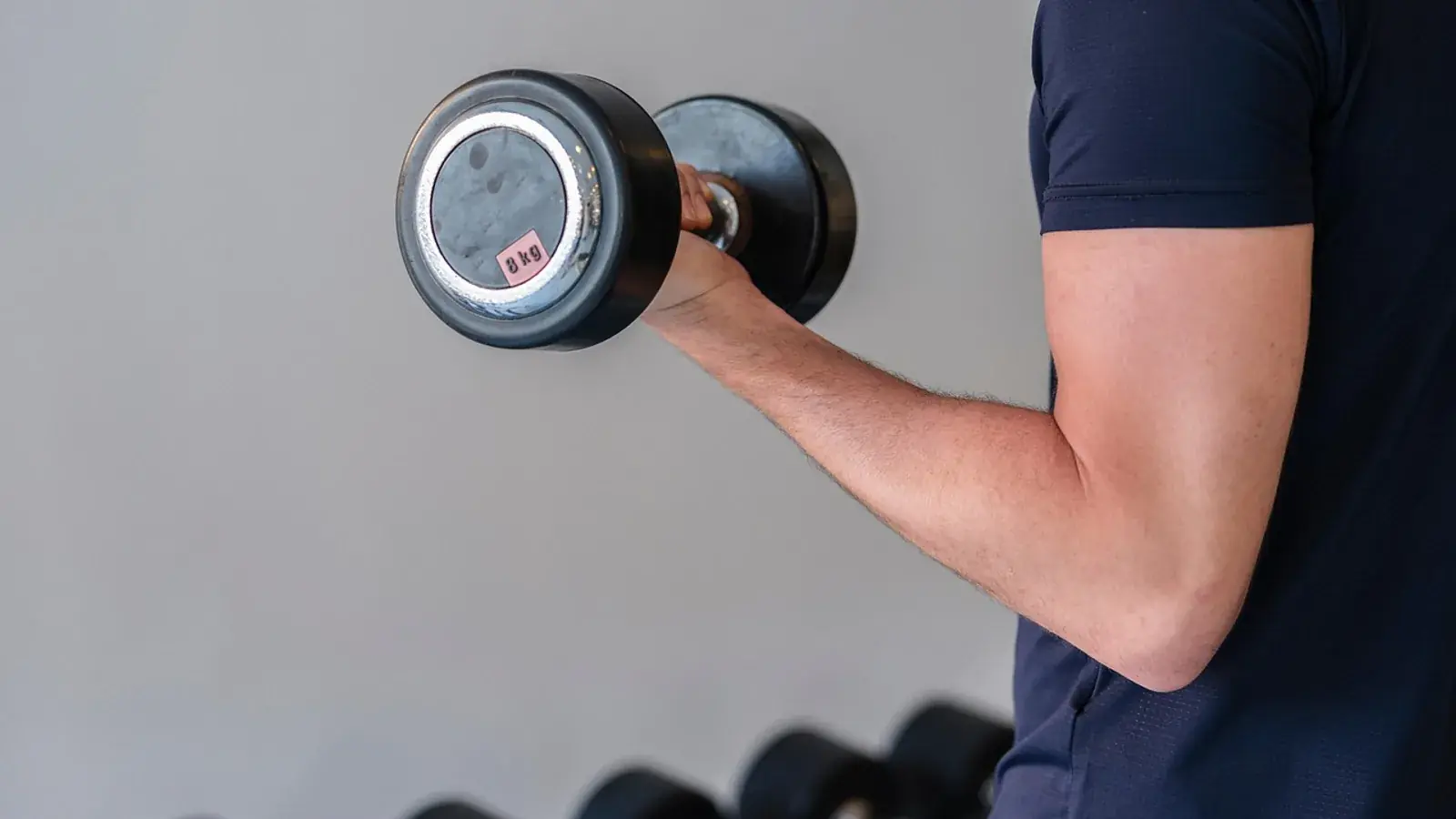 Arm eines Mannes im dunklen Sportshirt hebt eine Kurzhantel, darunter weitere Hanteln