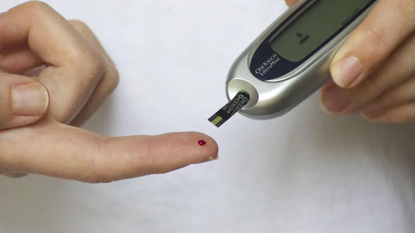 Blutstropfen an der Fingerkuppe, daneben ein Blutzuckermessgerät mit eingestecktem Teststreifen