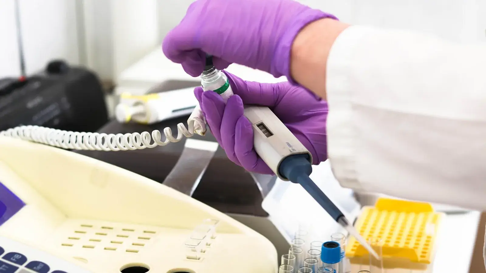 Hände in lila Handschuhen halten Pipette und Probenfläschchen an einem Laborgerät
